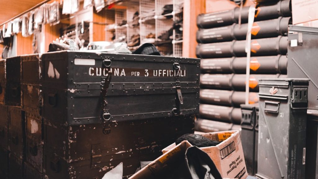 A busy military storage room filled with equipment and crates, labeled 'Cucina per 3 Ufficiali'.
